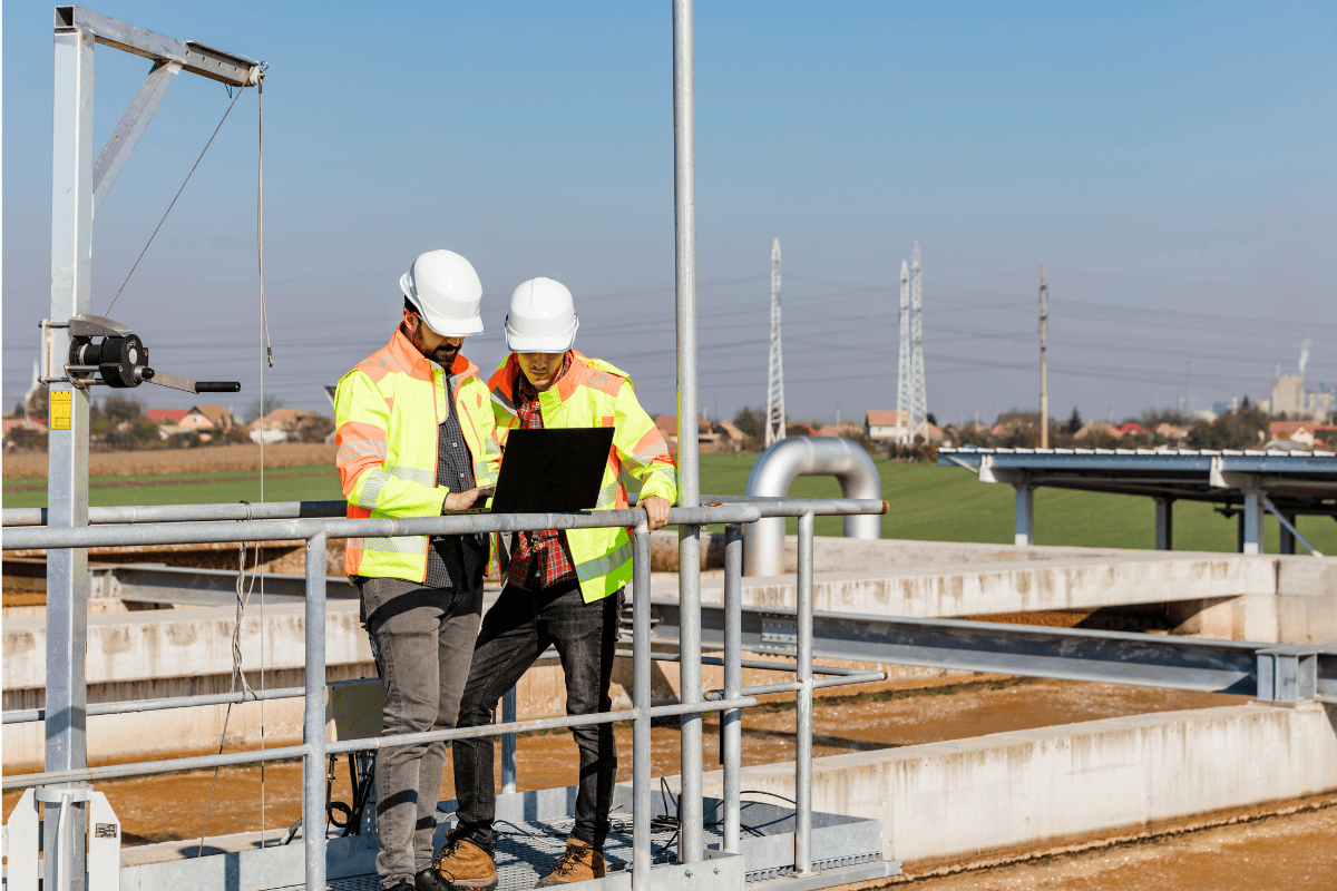 Outdoor photo of 2 men in hard hats and reflective vests looking at a laptop near a water facility.